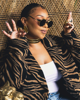 Tati Baker posing in Elorae's signature gold loop earrings, gold and silver halo rings and gold torque choker, with dark shades, sitting in a wicker chair with a cheeful expression.