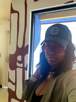 Elorae's customer Dara Segbefia posed in silver Metropolis Coil bangles and silver Torque choker with a green hat and green sweater against patterned wall.