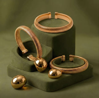 Elorae gold Icon Era Collection jewelry set including Halo rings and Metropolis Coil bangles on a green stand against a matching background.