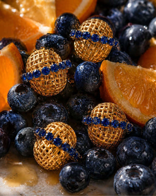 Four gold mesh-wrapped jewelry pieces with blue gemstone bands, arranged on a bed of blueberries and orange slices.