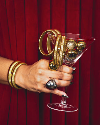 Elorae hand model posing with gold Metropolis Coil bangles and gold Halo rings holding a martini glass against a red curtain.