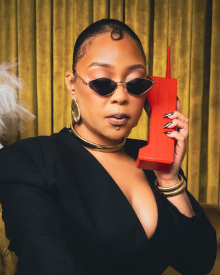 Tati Baker posing with red telephone in Elorae jewelry collection against a gold curtain.
