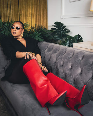Tati Baker posing in black outfit on grey couch wearing gold Elorae Icon Era Collection jewelry collection, red boots and black sunglasses.