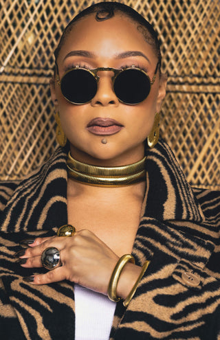 Tati Baker wearing Elorae's gold Metropolis Loop earrings, gold Torque choker set, gold Halo earrings and a zebra print coat with sunglasses against a textured background.