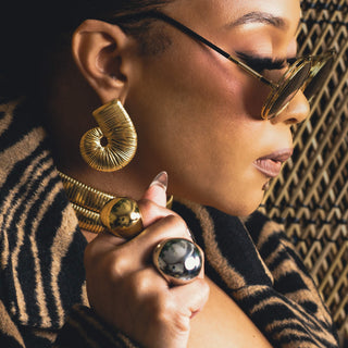 Tati Baker wearing wearing Metropolis gold earrings and sunglasses with a textured wall background