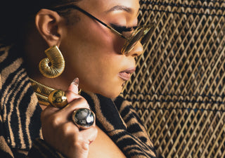 Tati Baker wearing wearing gold Metropolis Loop earrings, Halo rings, Coil Bangles, zebra jacket and sunglasses against a textured wall background.