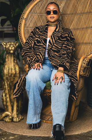 Tati Baker wearing wearing gold Metropolis Loop earrings, Coil Bangles  and sunglasses with a textured wall background.