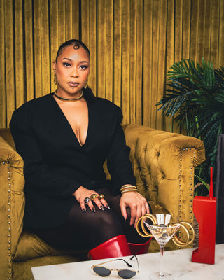 Elorae's founder Tati Baker dressed in black jacket and Elorae jewelry posed on a gold velvet couch, with marble table and red telephone.