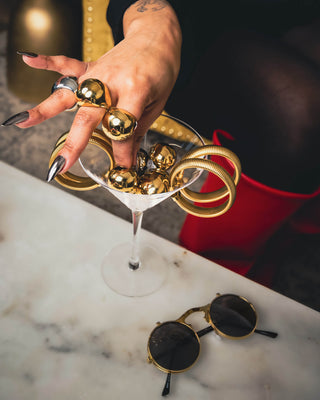 Hand holding a martini glass filled with Elorae's jewelry: Halo rings and Metropolis Coil bangles with sunglasses on marble surface